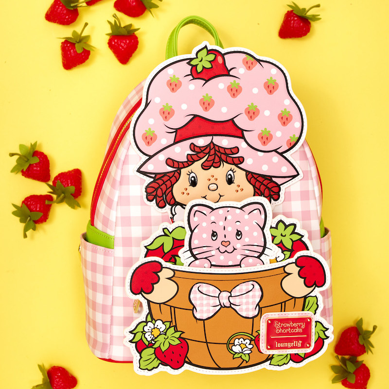 Exclusive Loungefly Strawberry Shortcake Custard Surprise Mini Backpack laying against a yellow background surrounded by strawberries. The bag includes light pink gingham print and Strawberry Shortcake as a large appliqué holding a basket of strawberries, Custard pops up from inside the basket.